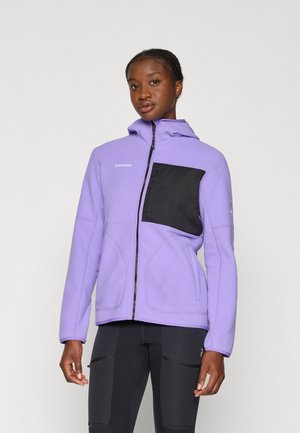 Personne vêtue d'une veste polaire violette avec une poche poitrail noire et un legging de sport noir, se tenant devant un fond clair uniforme.
