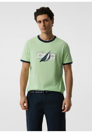 Mężczyzna z kręconymi włosami, ubrany w jasnozielony t-shirt z granatowym wykończeniem i żeglarskim logo, w połączeniu z ciemnymi spodniami i paskiem na jednolitym tle.