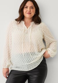 Kvinde med brunt hår iført en gennemsigtig, struktureret hvid bluse med snore og sorte læderbukser, stående med den ene hånd i lommen.