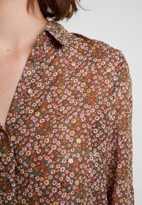 Brun blommig skjorta med små rosa, vita och guldblommor, långa ärmar och krage med knappar i brunt.