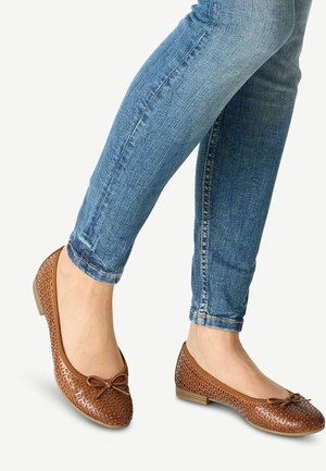 Brązowe skórzane baleriny z wyciętym kwiatowym wzorem, okrągłym noskiem, małym akcentem w postaci kokardy oraz niskim blokowym obcasem, połączone z niebieskimi skinny jeansami.