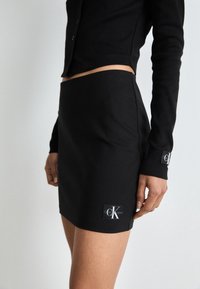 Femme portant une mini-jupe noire taille haute et un top noir à manches longues, tous deux avec des écussons du logo Calvin Klein Jeans.