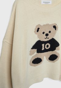 Pull en tricot couleur crème avec un grand motif d'ourson portant un t-shirt noir affichant le numéro 10. Encolure et ourlet côtelés.