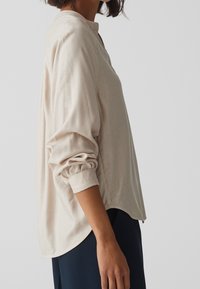 Blusa a maniche lunghe beige con scollo rotondo, maniche arricciate e orlo curvo. Tessuto liscio, leggero e vestibilità comoda.