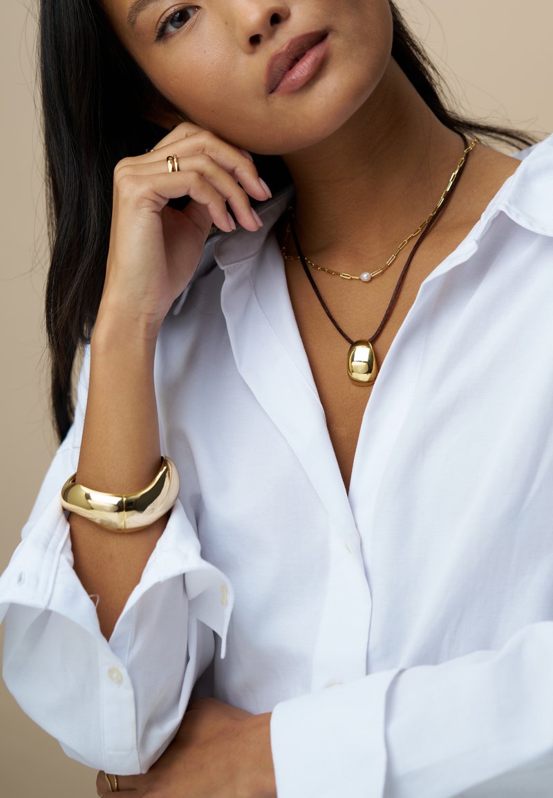 Collier pendentif en or sur un cordon marron, superposé avec une chaîne en or à maillons et une petite perle. Le modèle porte une chemise blanche boutonnée et un bracelet jonc en or.