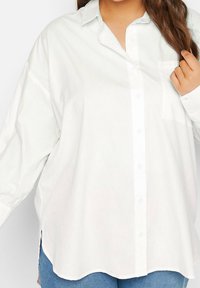 Vit skorta med krage, långa ärmar och bröstficka; har rundad nederkant och sidventiler för enkel rörlighet.