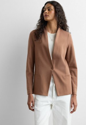 Femme aux cheveux bouclés portant un blazer marron à un seul bouton sur une chemise blanche et un pantalon blanc, sur un fond gris uni.