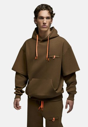 Jeune homme portant un sweat à capuche marron oversize et un pantalon assorti avec des cordons orange et de petites poches avant, debout devant un fond uni.