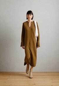 Abito lungo in maglia verde oliva con scollatura a V, maniche lunghe e spacchi laterali, abbinato a stivali alla caviglia beige e una borsa a tracolla bianca.
