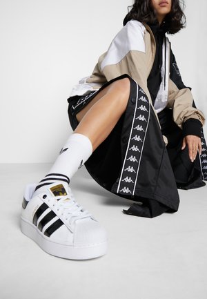 Persona con chaqueta beige y blanca, pantalones deportivos negros con rayas blancas con el logo, zapatillas Adidas blancas y calcetines blancos, sentada en el suelo.