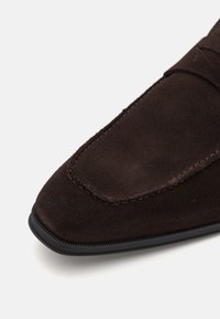 Barna velúr slip-on cipő, lekerekített orral, kontrasztos díszítő varrással és fekete gumitalppal. Sima felület, enyhe fényességgel.