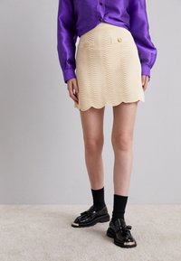 Mini-jupe couleur crème avec un ourlet festonné, motif en vagues et deux poches avant ornées de boutons dorés, assortie à des chaussures et des chaussettes noires.