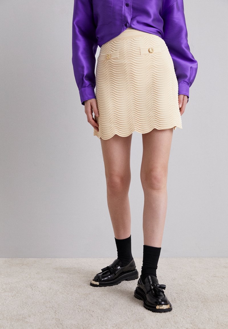 Mini-jupe couleur crème avec un ourlet festonné, motif en vagues et deux poches avant ornées de boutons dorés, assortie à des chaussures et des chaussettes noires.