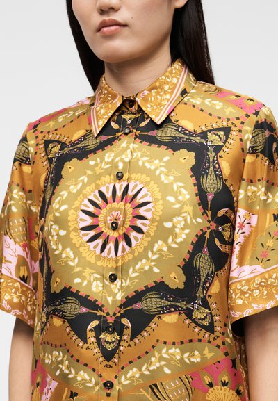Short-sleeve shirt in gold fabric featuring intricate floral and abstract patterns in black, pink, and green, with a collared neckline and buttons.