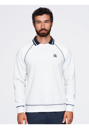 Homme portant un polo blanc à manches longues avec un col bleu marine et un petit emblème brodé sur la poitrine gauche, debout devant un fond blanc uni.