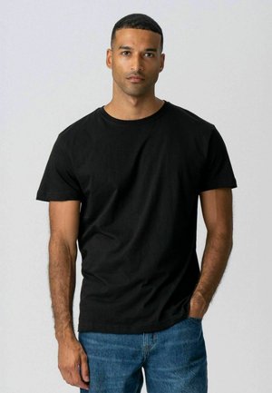 Noor mees lühikeste juustega, kandes musta t-särki ja siniseid teksaseid, seisab ühe käega taskus ühevärvilise tausta ees.