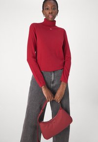 Maglione a collo alto rosso realizzato in tessuto liscio, abbinato a jeans grigi a gamba larga a vita alta. Tiene in mano una piccola borsa rossa con cerniera.