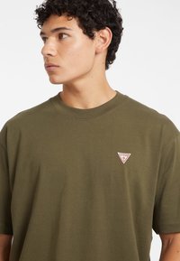 Tricou din bumbac verde măsliniu, cu guler tip crew și o mică etichetă logo triunghiulară roșie și albă pe partea stângă a pieptului.