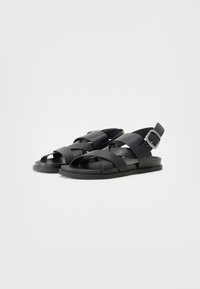 Sorte sandaler laget av syntetisk materiale med kryssende stropper, åpen tå-design og en sideknepping for justering. Flat gummisåle.
