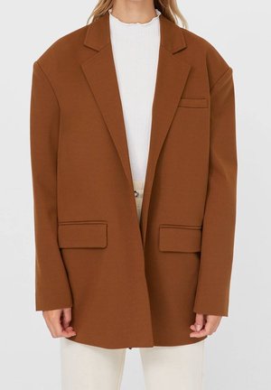 Vrouw die een oversized bruine blazer draagt met notch-revers, twee klepzakken aan de voorkant, een borstzak en lange mouwen over een wit topje.