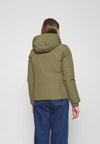 Olive green puffer jacket with a hood, featuring a fitted design, quilted texture, and elastic cuffs. Paired with blue wide-leg jeans.