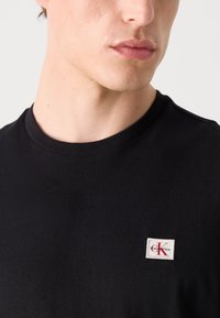 Približna snimka osobe koja nosi crnu majicu s okruglim izrezom i malom bijelo-crvenom oznakom Calvin Klein na prsima.