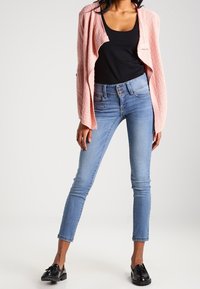 Heller rosa, strukturierter Cardigan über einem schwarzen Tanktop; kombiniert mit schlanken, hellblauen Jeans und schwarzen Slippern.