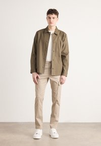 Olivgrön knäppjacka med en enda bröstficka, i kombination med beige cargobyxor och vita sneakers, allt visat mot en neutral bakgrund.