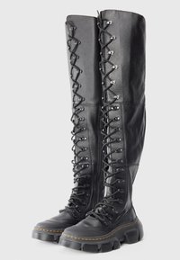 Bottes montantes en cuir noir à lacets avec semelles épaisses et robustes, et coutures jaunes autour de la base, se tenant côte à côte sur un fond blanc.