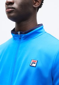 Μπλε ζακέτα με φερμουάρ κατασκευασμένη από ομαλό υλικό, με κοντράστ ναυτικό γιακά και μικρό λογότυπο FILA στο στήθος.