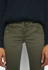 Person klädd i olivgröna byxor med dubbel knappstängning och en mörkblå tröja, med händerna avslappnat vid sidorna.