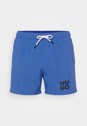 Zilā peldbikses vieglā audumā ar elastīgu jostasvietu un balto saiti. Apakšējā labajā stūrī ir melns "HUGO" logo.