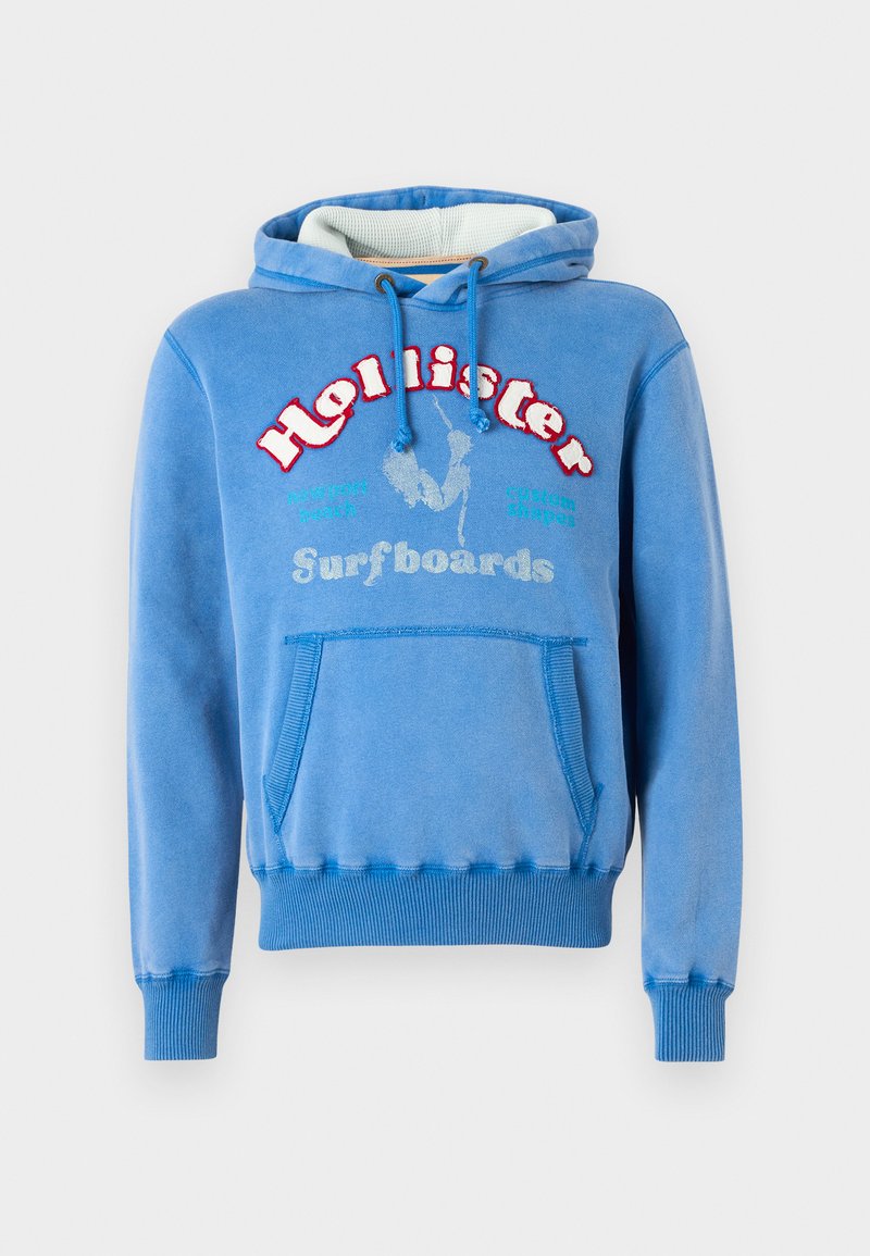 Sudadera azul con puños y dobladillo acanalados, un bolsillo frontal y un logo bordado de "Hollister Surfboards" en rojo y blanco. Textura suave.