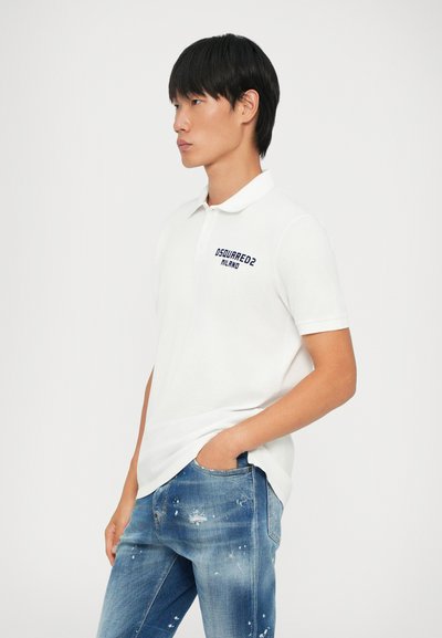 Fehér póló ing, hajtott gallérral és sötétkék hímzett felirattal a bal mellrészen. A modellen világoskék, megkopott farmernadrág van.