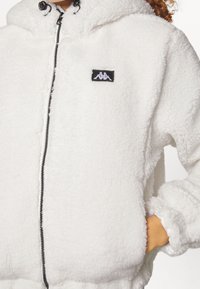 Chaqueta de forro polar blanca con capucha, cierre de cremallera y detalles en negro. Presenta un pequeño logo y puños elásticos para mayor comodidad.