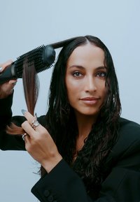 Vrouw met donker haar die een ronde elektrische haarborstel gebruikt om een sectie nat haar te stylen tegen een effen lichte achtergrond.