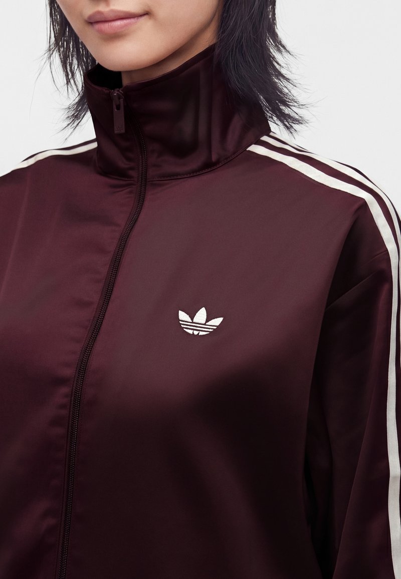 Gros plan d’une personne portant une veste zippée bordeaux avec des bandes blanches sur les épaules et un petit logo blanc sur la poitrine.