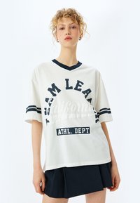 Hvid bomulds T-shirt med navy V-hals, der har fed sort tekst "TEAM LEARN" og "ATHL. DEPT," akkompagneret af stribede ærmer.
