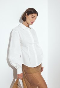 Witte button-up shirt met volumineuze mouwen, voorzien van een puntige kraag en een gestructureerde stof; gecombineerd met een doorzichtig beige rokje en een bruine tas.