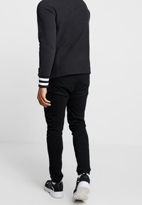 Felpa nera con polsini a righe bianche, pantaloni neri affusolati e sneakers nere, che mostrano un tessuto testurizzato e un design aderente sul retro.