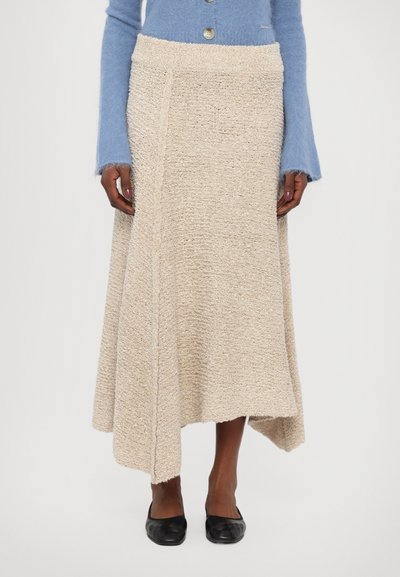 Jupe beige texturée avec un ourlet asymétrique, fabriquée à partir d'un matériau tissé. Associée à un pull bleu clair et des ballerines noires.