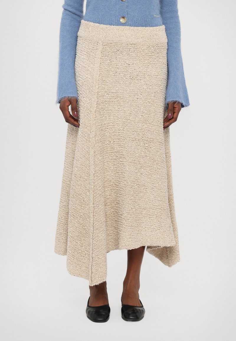 Beige textured skirt with an asymmetrical hem, made of a woven material. Paired with a light blue sweater and black flats.
