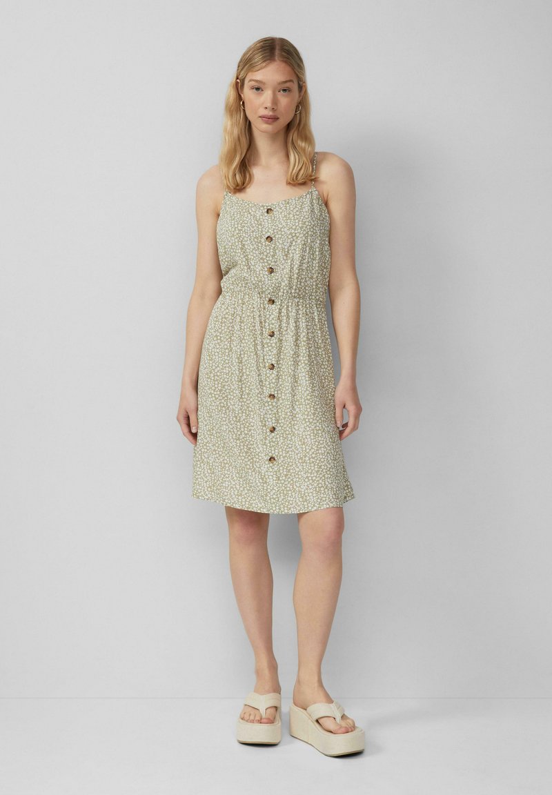 Grünes Blumenkleid mit tailliertem Schnitt, Knopfverschluss vorne und Spaghettiträgern. Getragen mit leichten Plateau-Sandalen. Weicher Stoff und lockere Passform.