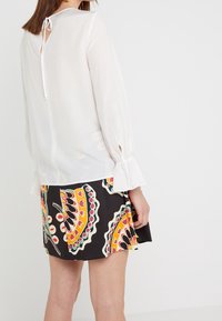 Vit lätt blus med långa ärmar, med en knytning i ryggen. Matchas med en svart kjol prydd med ett färgglatt paisleymönster.