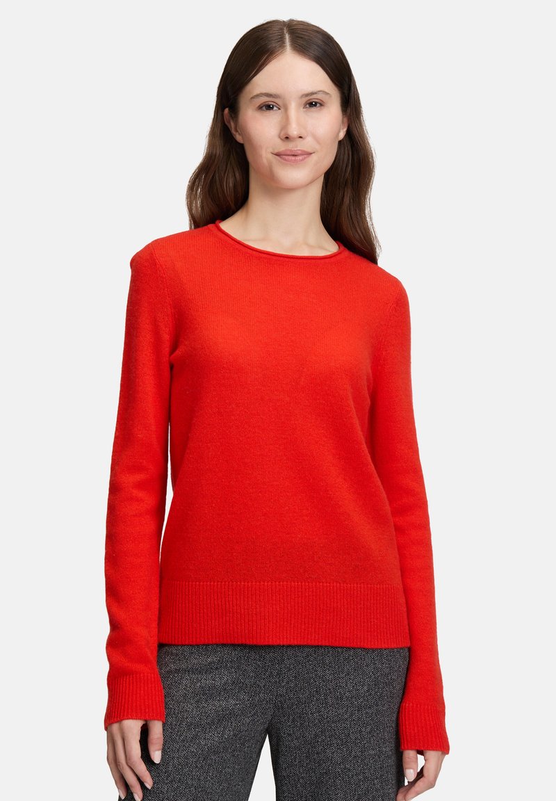 Frau mit langen braunen Haaren, die einen leuchtend roten langärmeligen Pullover und graue gemusterte Hosen trägt, steht vor einem weißen Hintergrund.