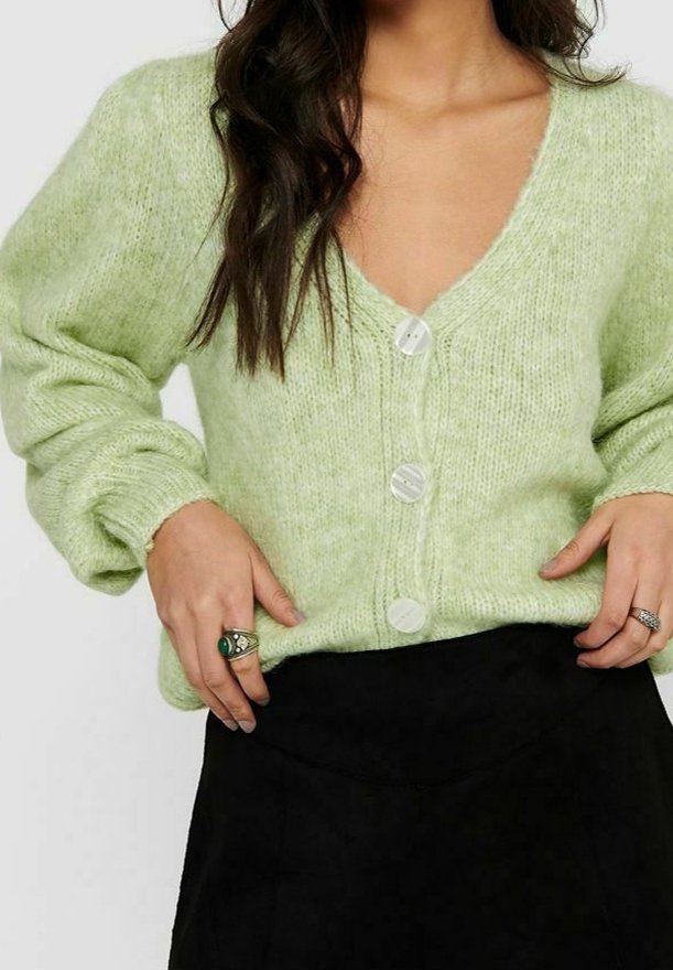 Hellgrüne gestrickte Strickjacke mit tiefem V-Ausschnitt, vier weißen Knöpfen und Ballonärmeln. Wird über einem schwarzen Kleidungsstück getragen.