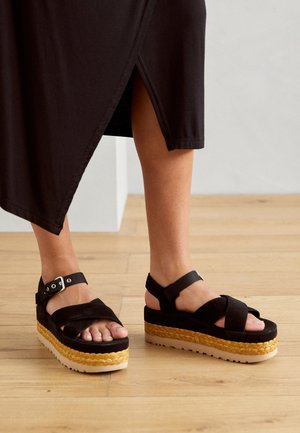 Pieds portant des sandales compensées noires avec une semelle intermédiaire tannée tissée et des brides à la cheville, assortis à une jupe noire fendue à l'avant, debout sur un sol en bois.
