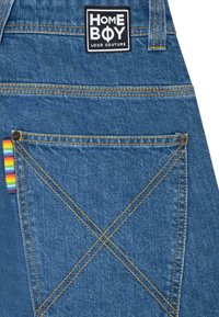 Mørkeblå denim overaller med gule sømmer og en lapp som leser "HOME BOY LOUD COUTURE." Har en regnbue-stripet detalj på baklommen.
