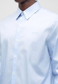 Chemise bleu clair boutonnée portée par une personne, avec un col pointu et un nom de marque brodé discrètement sur la poitrine.