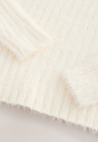 Pull en maille couleur crème avec une texture douce et duveteuse, présentant des détails côtelés sur les manches et le corps.